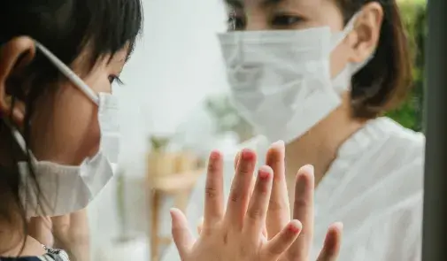 woman in mask holds up hand to a child in mask who is on the other side of a window