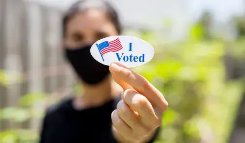 woman in coronavirus mask holds a sticker that says quote i voted unquote