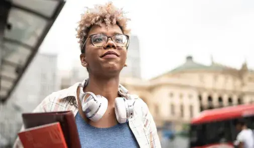 black woman with glasses holds books, looks toward the sky, has headphones resting around her neck