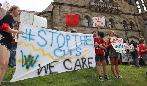 West Virginia University students, faculty, and employees holding signs. The sign in the foreground reads "#Stop the Cuts. We Care."