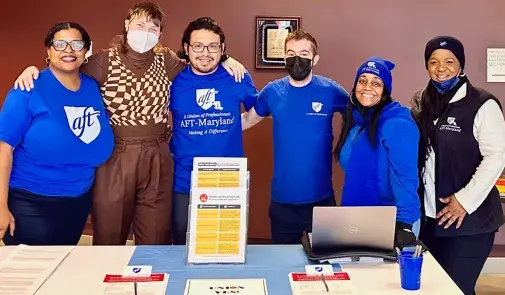 six adults behind a table covered in handouts. the adults are smiling with their arms around each other as they pose for the picture