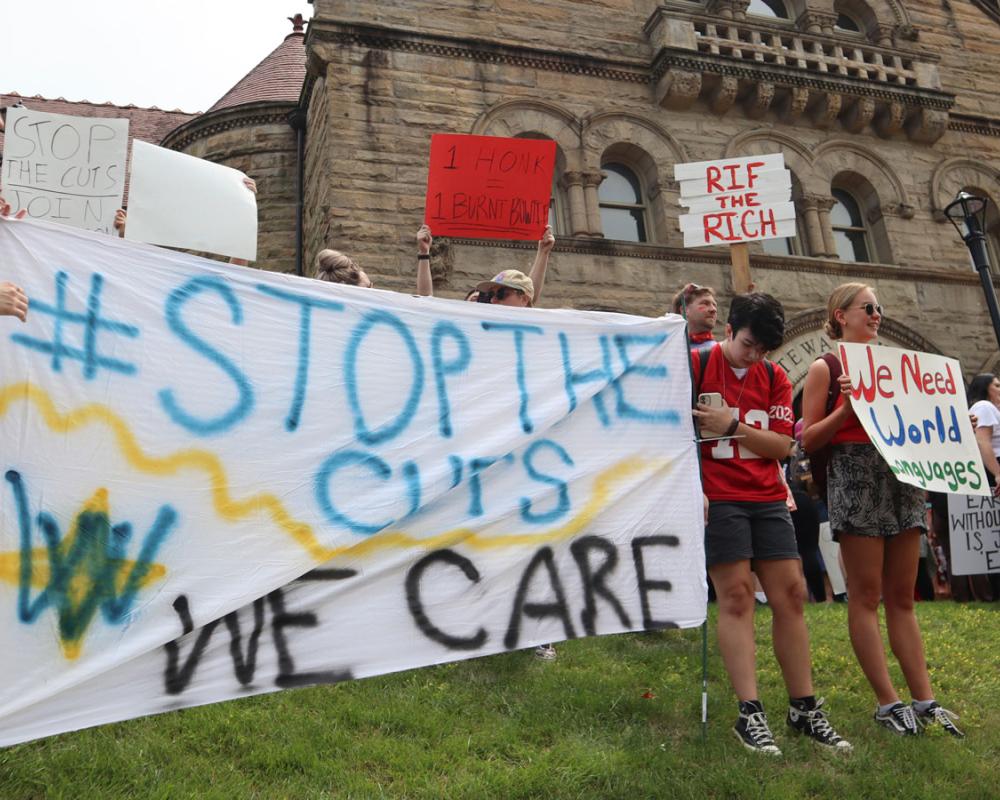 West Virginia University students, faculty, and employees holding signs. The sign in the foreground reads "#Stop the Cuts. We Care."