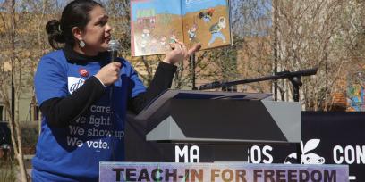 Montserrat Garibay, wearing an AFT tshirt, holds a picture book as she reads aloud into a microphone onstage at a teach-in for freedom event.