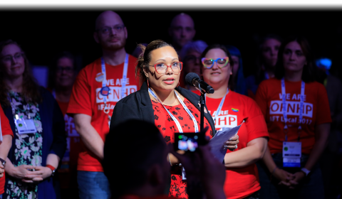 AFT member speaks at Convention 2024 with members behind wearing shirts that read "We are OFNHP"